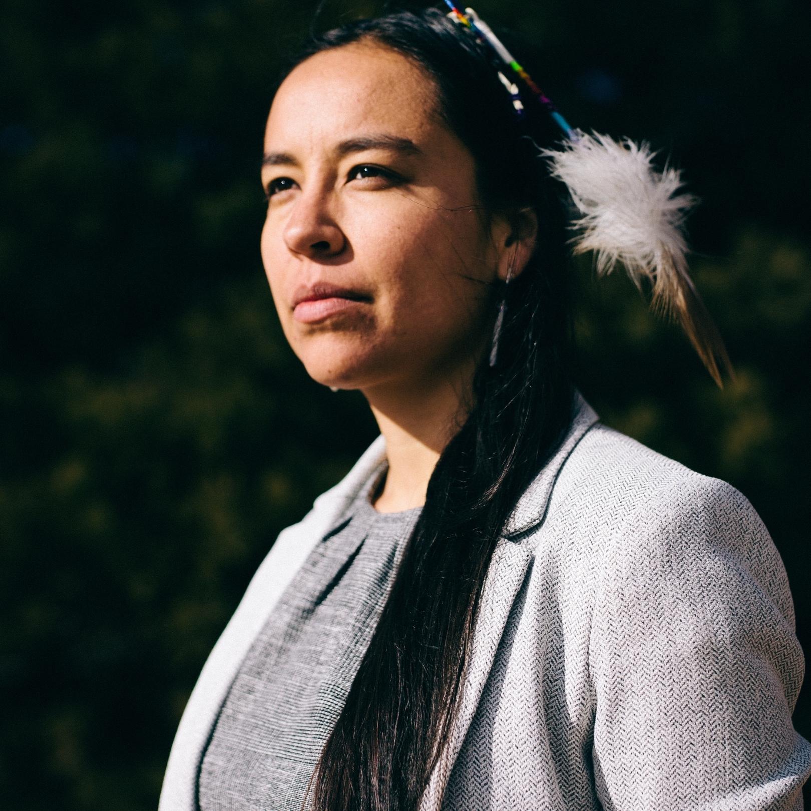 A picture of a Native American woman, Lyla June, in a gray business suit with a feather in her hair.